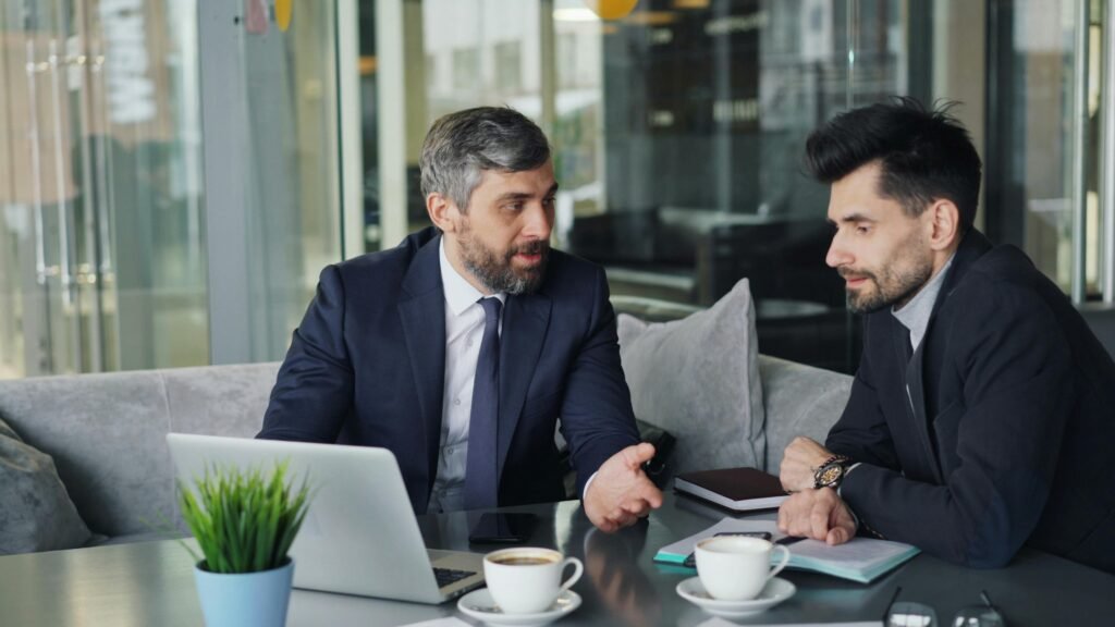 Two businessmen in a café having a professional discussion with coffee and a laptop.